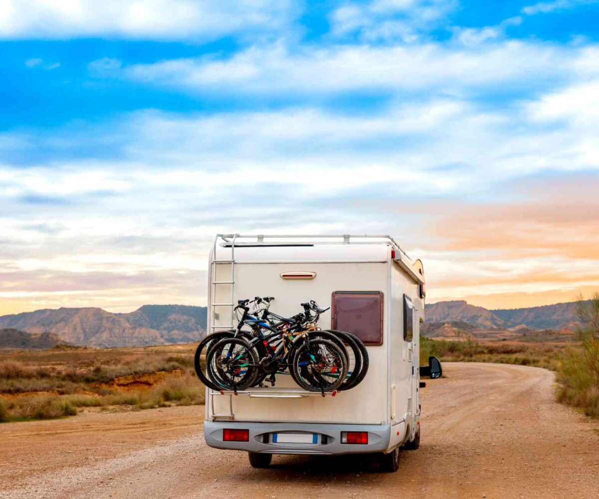 A camper van carrying bikes driving along a desert dirt road at sunset.