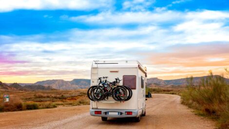 A camper van carrying bikes driving along a desert dirt road at sunset.