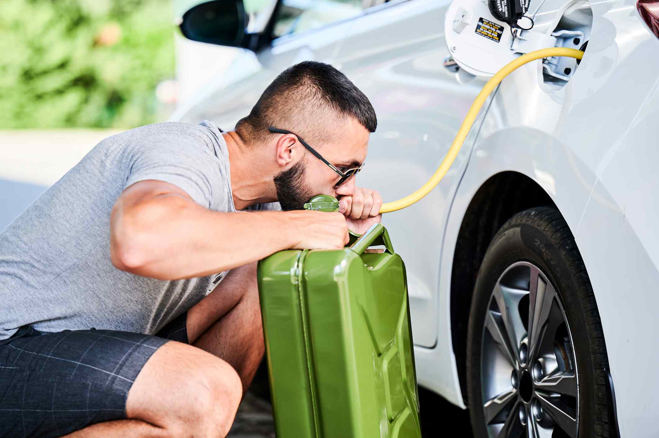 A man siphons gas from his car into a canister on his driveway, sucking on the hose.