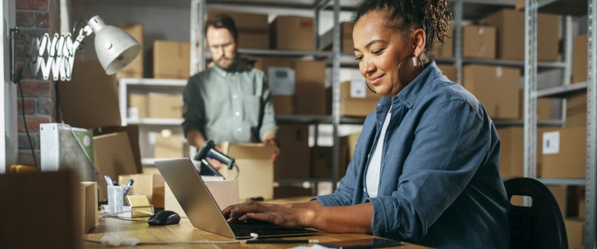 A smiling woman works on a laptop while a coworker packs boxes on shelves.