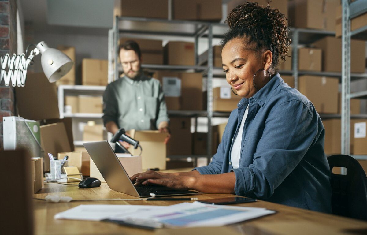 A smiling woman works on a laptop while a coworker packs boxes on shelves.
